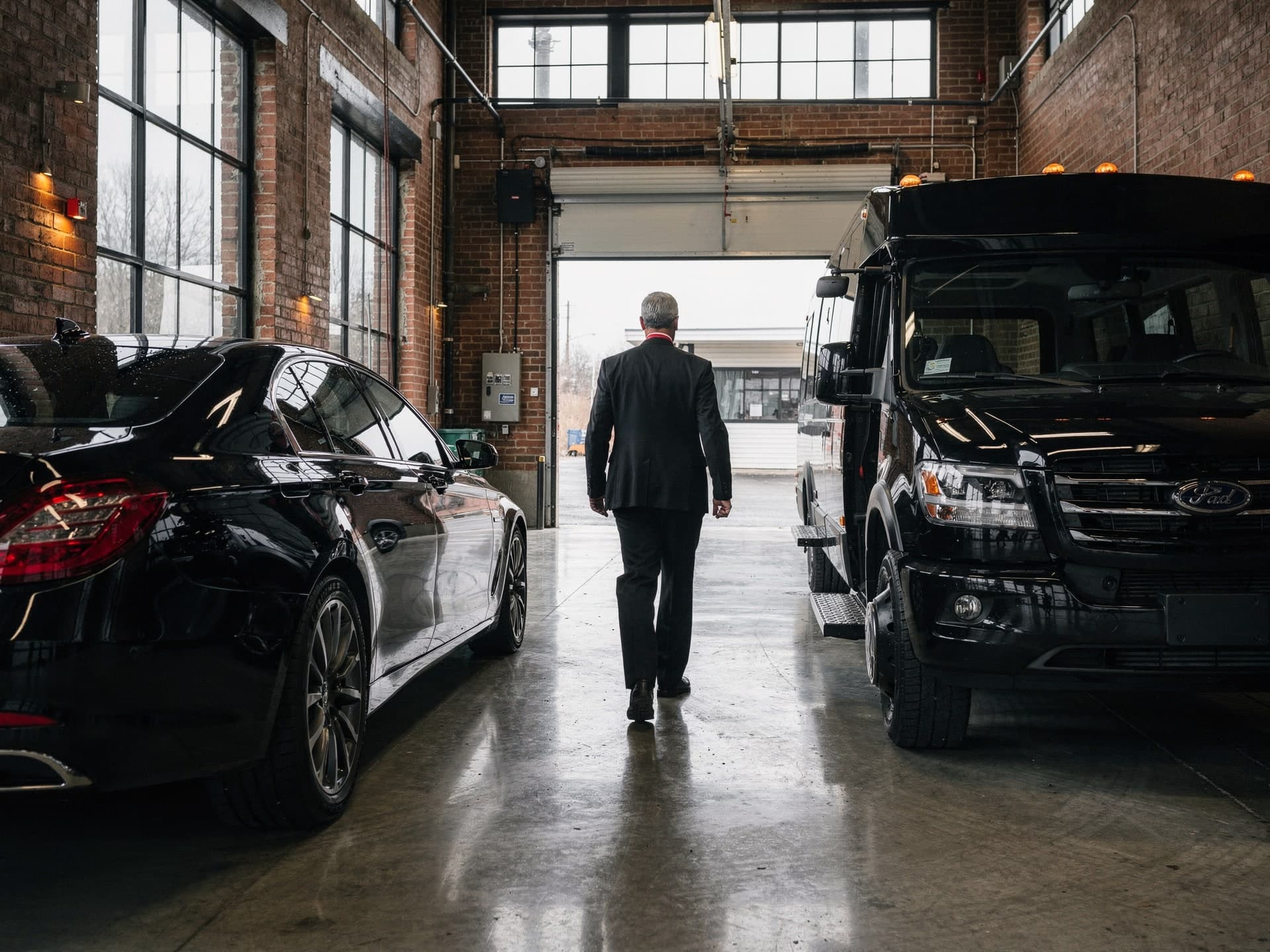 Seen from inside the Methuen garage, a chauffeur in a black suit walks away between a black executive sedan and a black shuttle bus toward the open roll-up door and the daylight beyond.