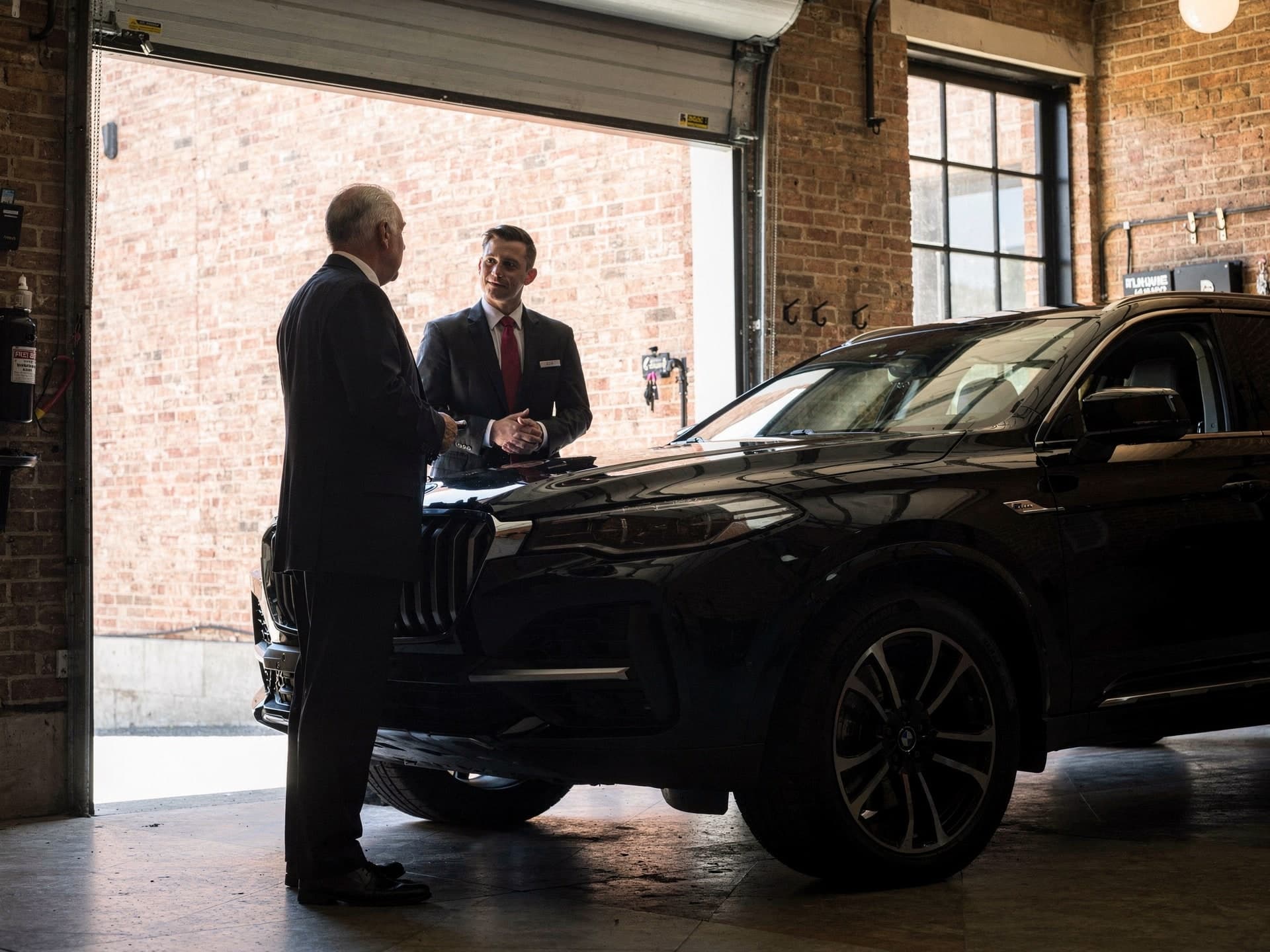 Two chauffeurs in dark suits stand talking at the front wing of a black SUV inside a brick garage bay, the older man with his back to the camera and the younger one facing him in a red tie, daylight coming in through the raised roll-up door behind them.