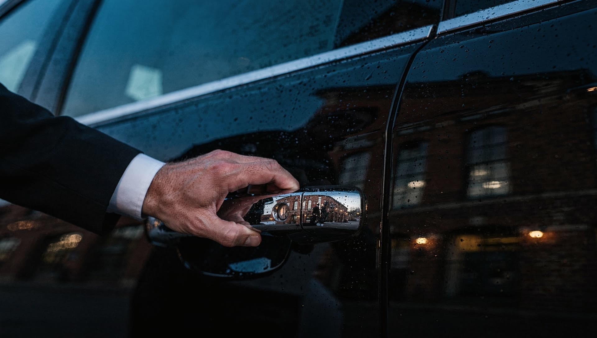 A chauffeur's hand resting on the polished chrome handle of a black SUV at dusk, water beading on the dark paint and the reflection of a brick mill building sharp in the door.