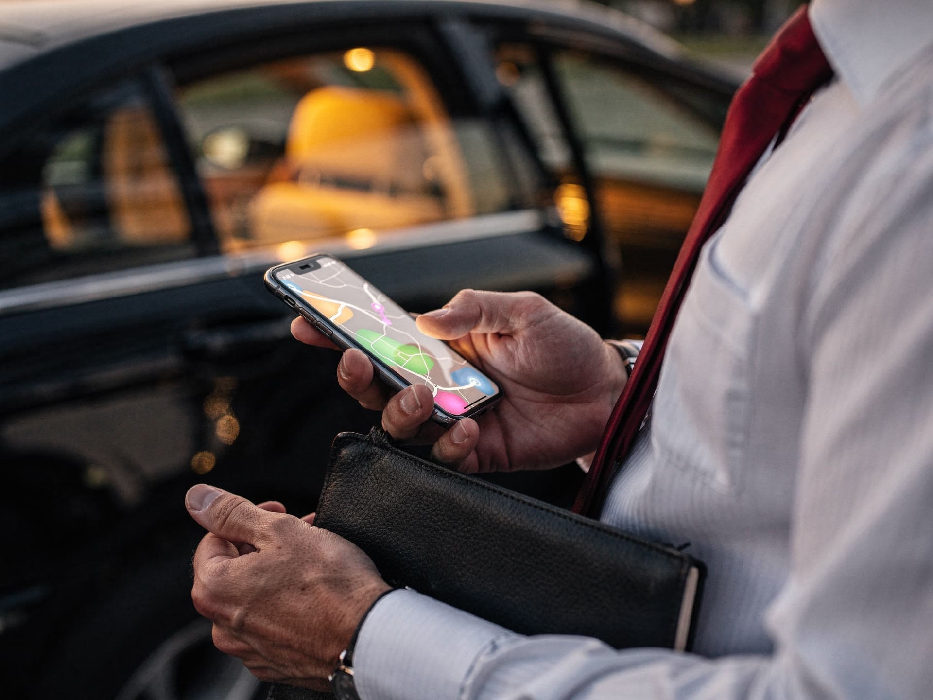 Close on the hands of a chauffeur in a white shirt and red tie holding a phone that shows a route map, a leather-bound notebook and a black sedan's door open behind him in warm evening light.