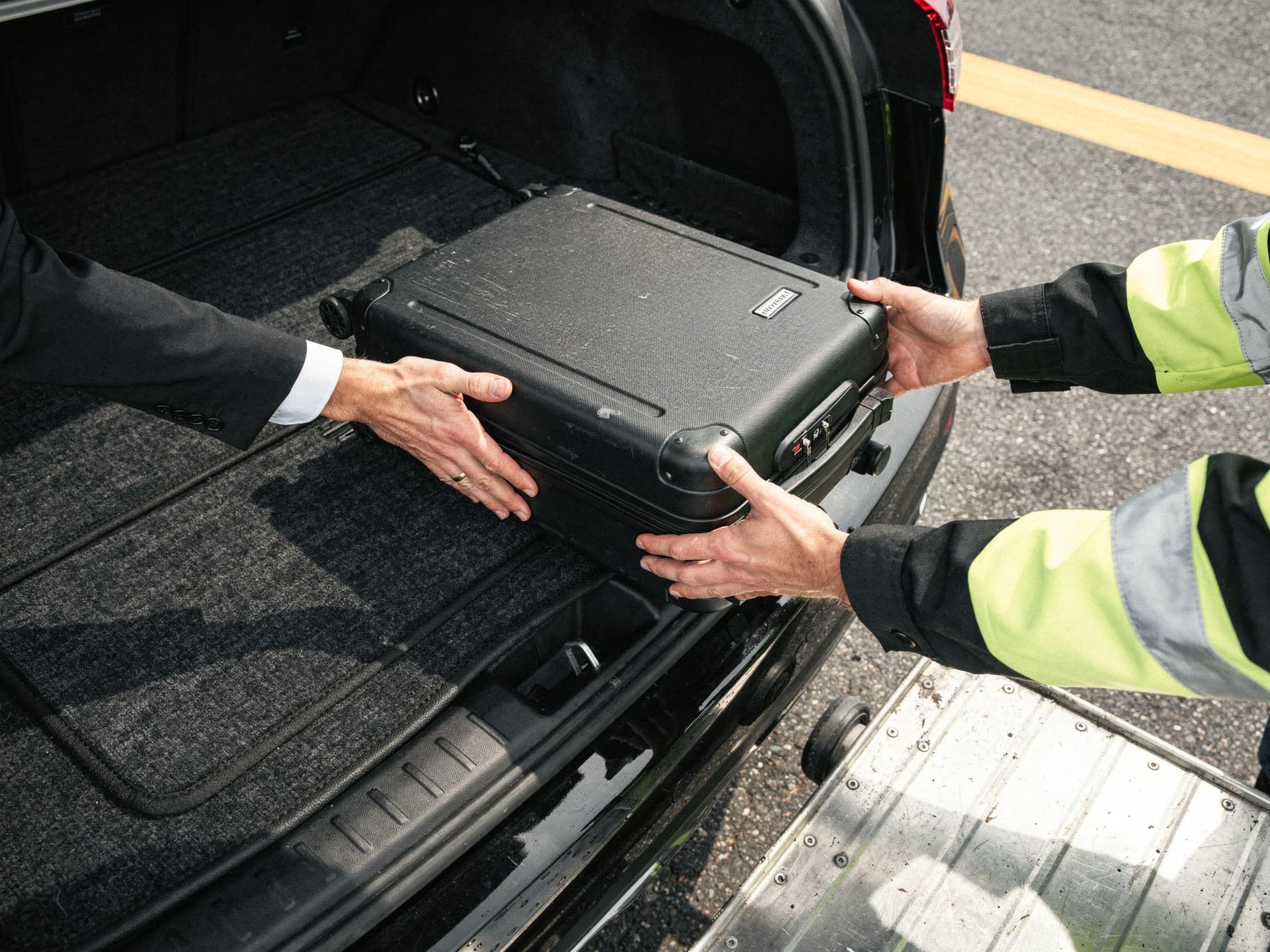 Hands and forearms moving suitcases from a ramp cart into an open car trunk on apron tarmac, painted apron lines in the frame.