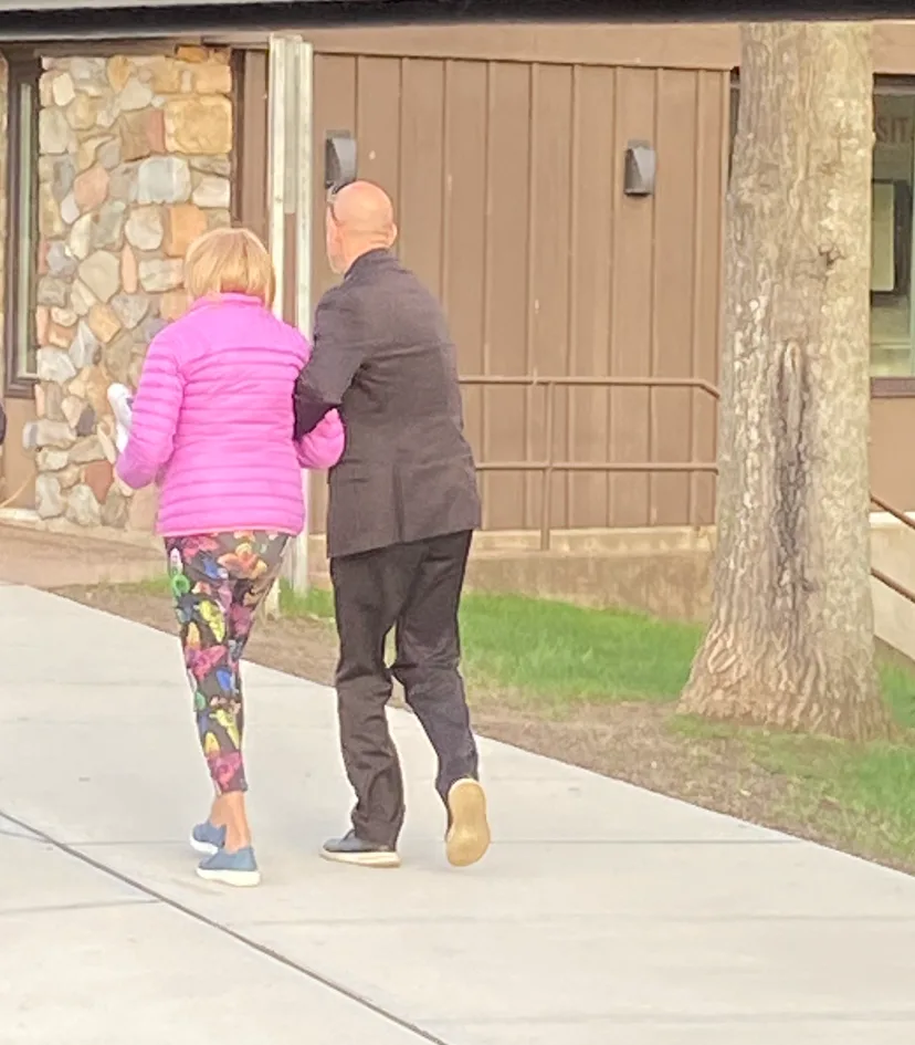 A chauffeur in a dark suit walks arm in arm with a woman in a pink jacket along a sidewalk beside a stone building