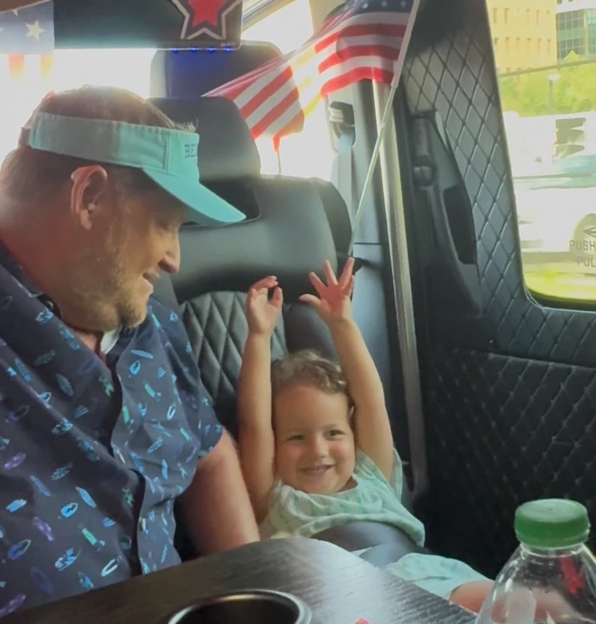 A man in a mint visor grins at a toddler with both arms raised on the rear bench of a party bus, small American flags above the window
