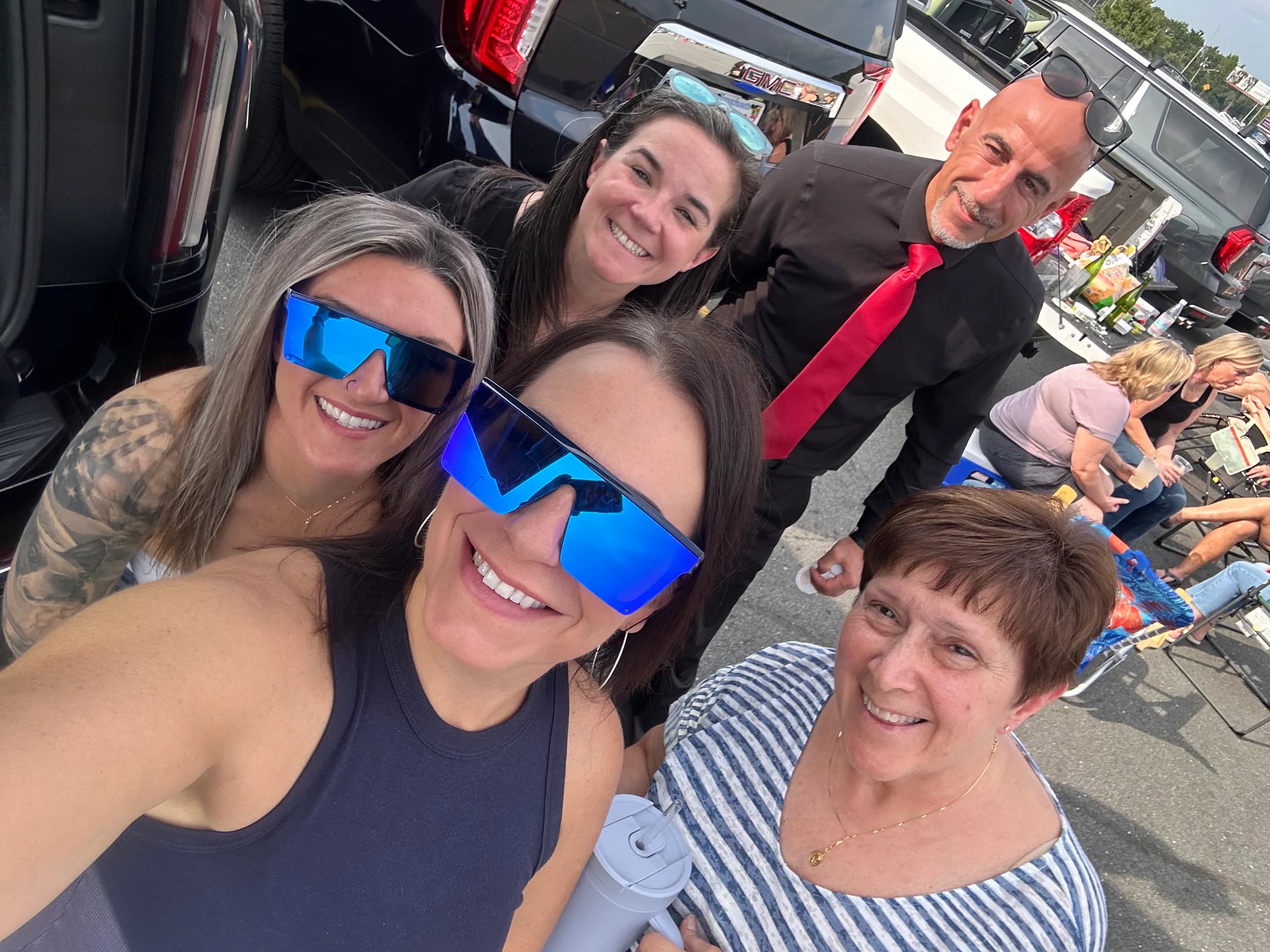 Four women smiling in a parking lot beside a black GMC SUV, a chauffeur in a black shirt and red tie behind them