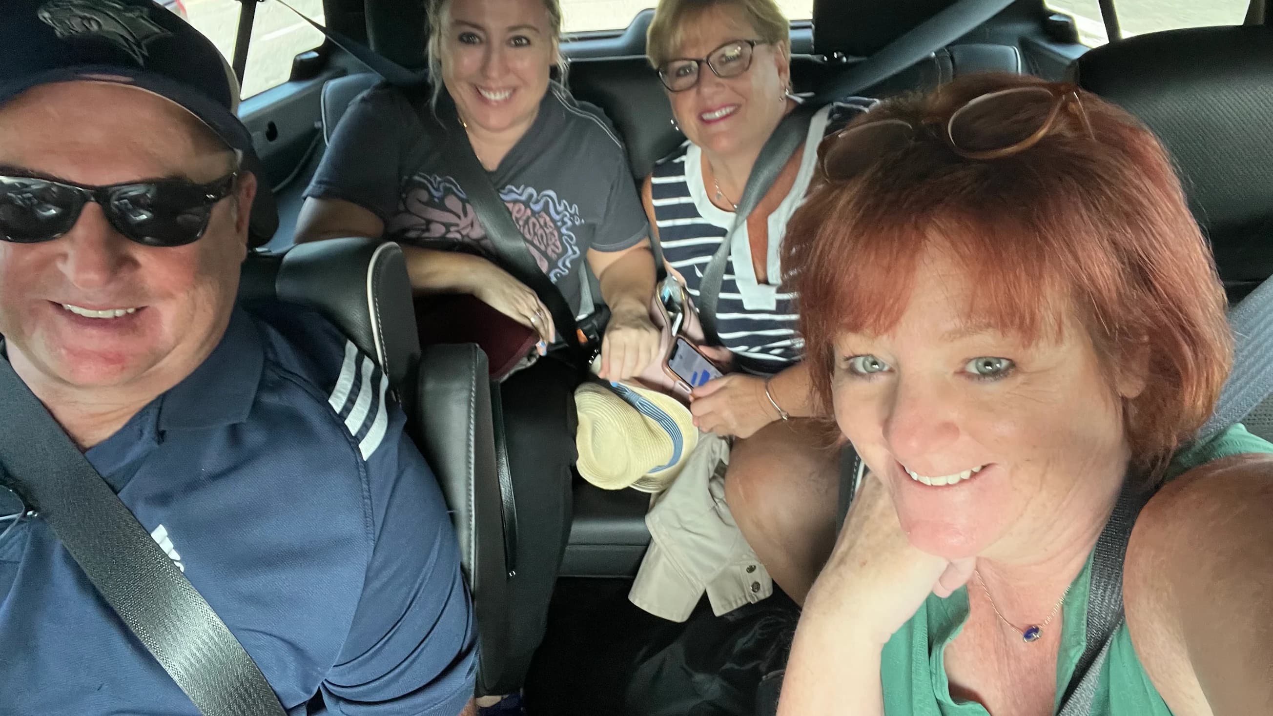 Four passengers buckled into the rear seats of an SUV, all smiling at the camera, a straw sun hat on one lap