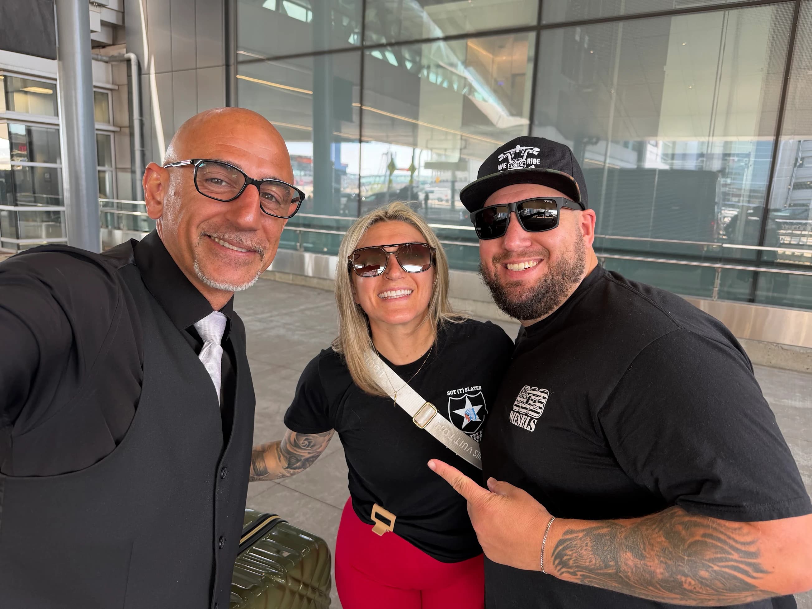 A chauffeur in a black vest and silver tie takes a selfie with a smiling couple at an airport terminal entrance