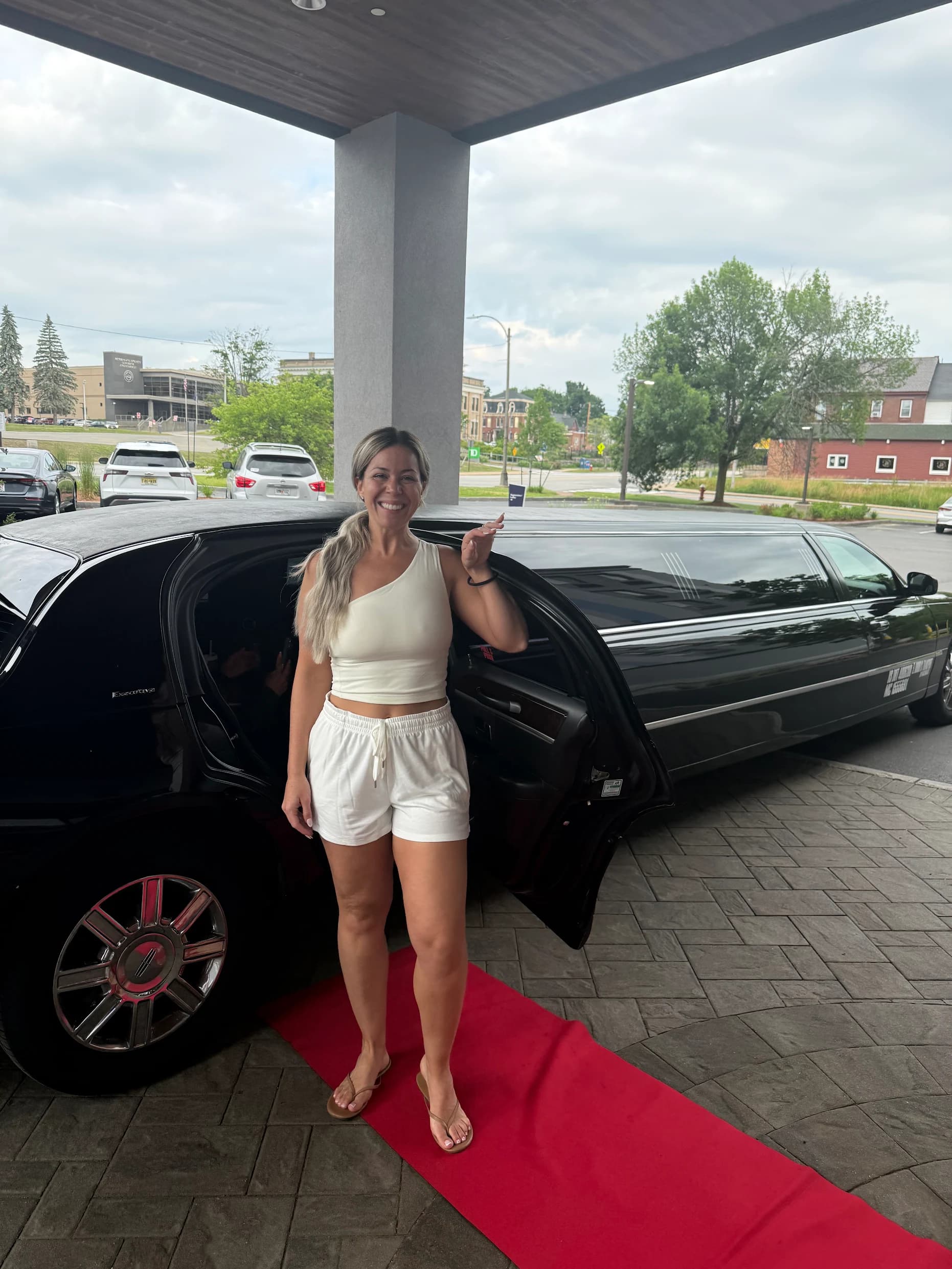 A woman in white smiling on a red carpet beside the open door of a black stretch limousine under a hotel portico