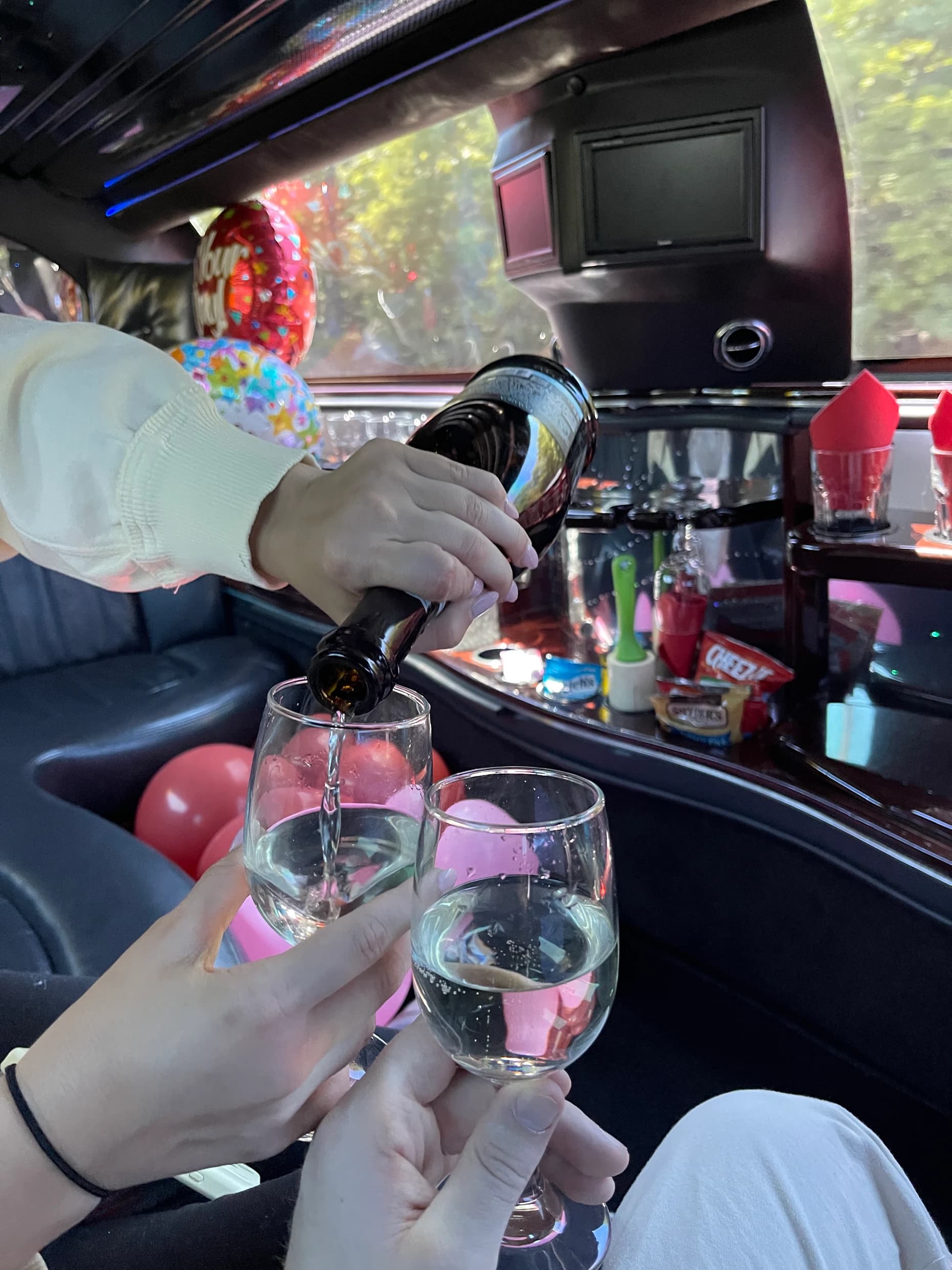 Sparkling wine being poured into two glasses inside a limousine, foil balloons and a stocked snack bar behind