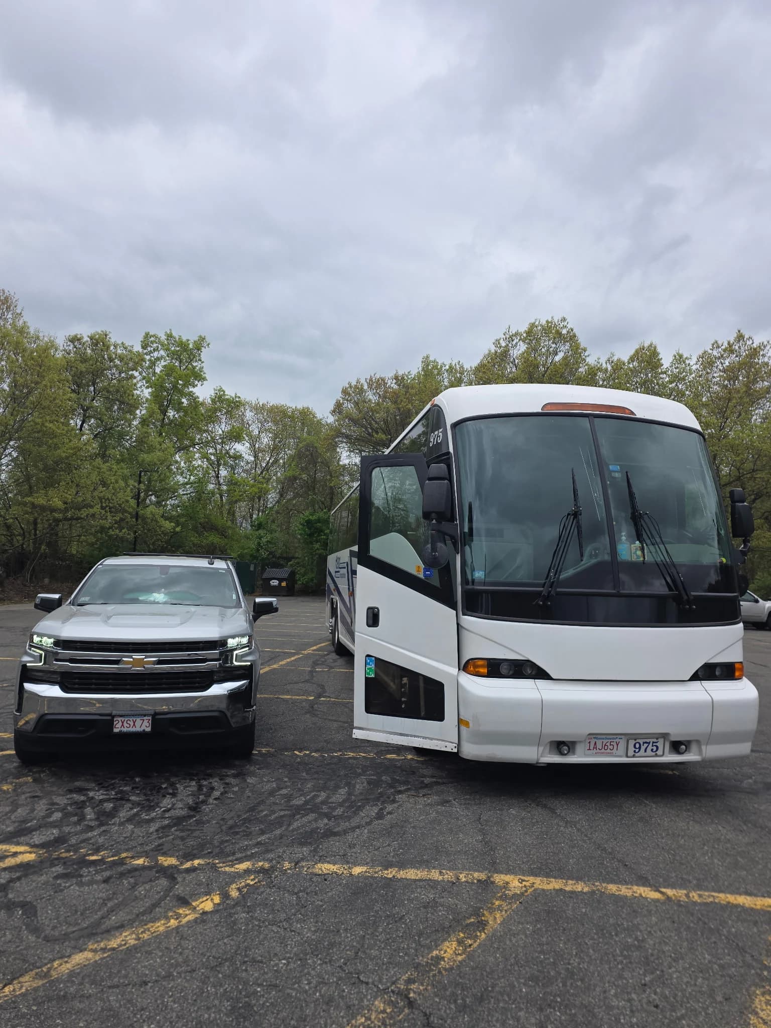 A white motor coach with its door open parked beside a silver Chevrolet pickup in a lot under an overcast sky