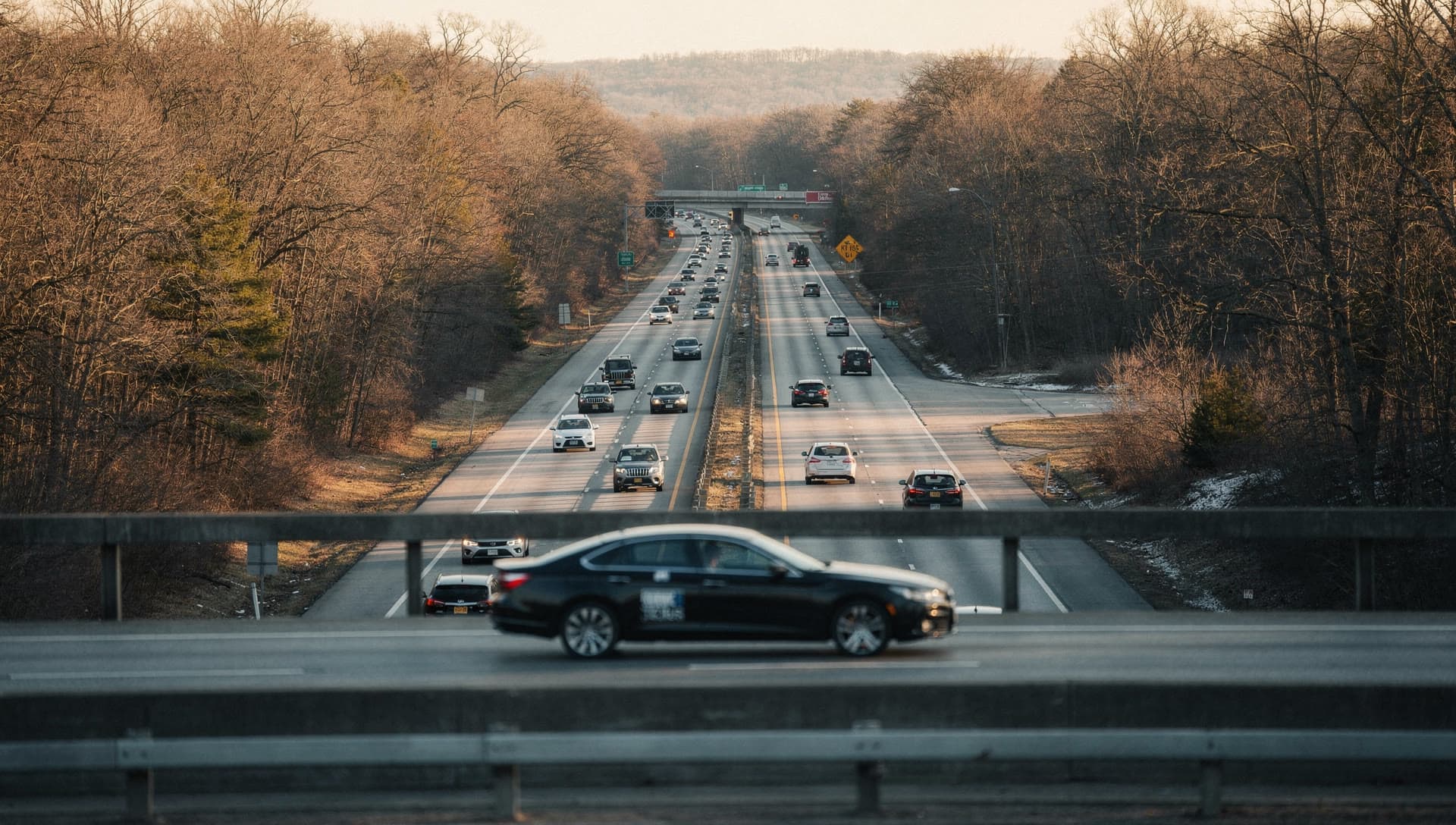 An elevated view of a northbound divided highway running through New England hardwoods at golden hour with light traffic on it.
