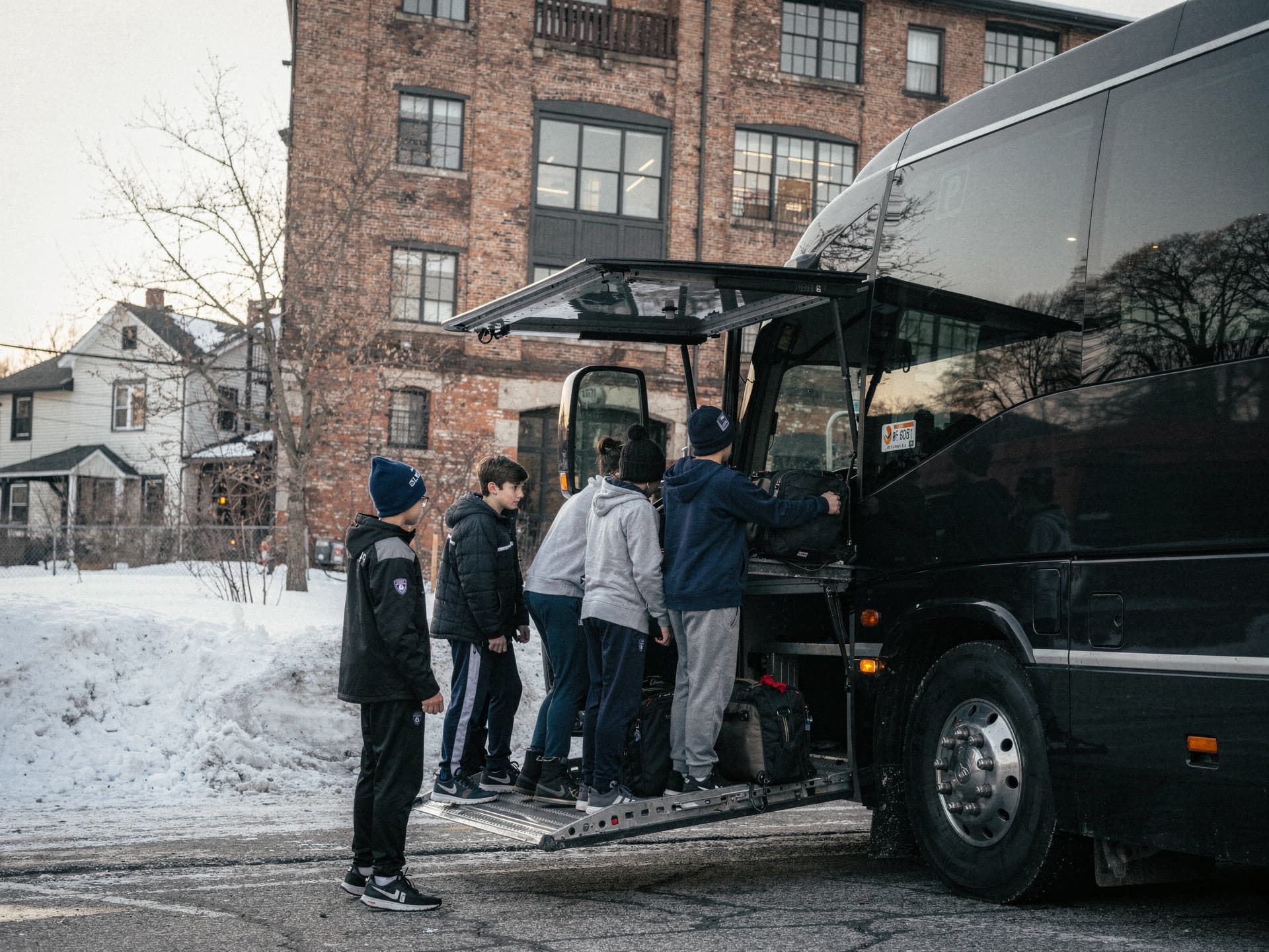 A youth sports team loading bags into the open under floor luggage bay of a coach in early morning light while an adult coach counts them on.