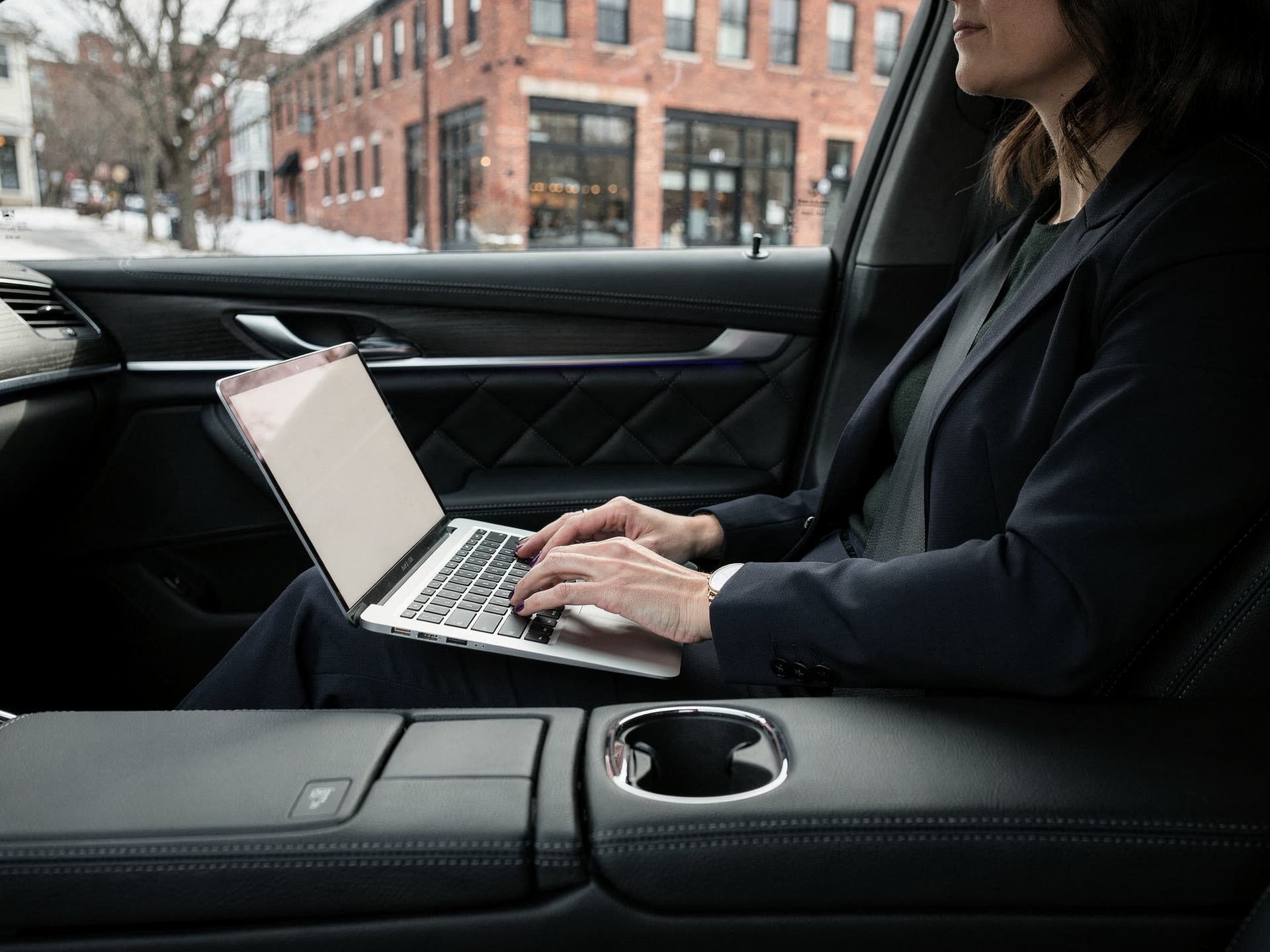 The rear cabin of an executive sedan seen from the opposite seat, a laptop open on a lap and window light raking across it as the city moves past.