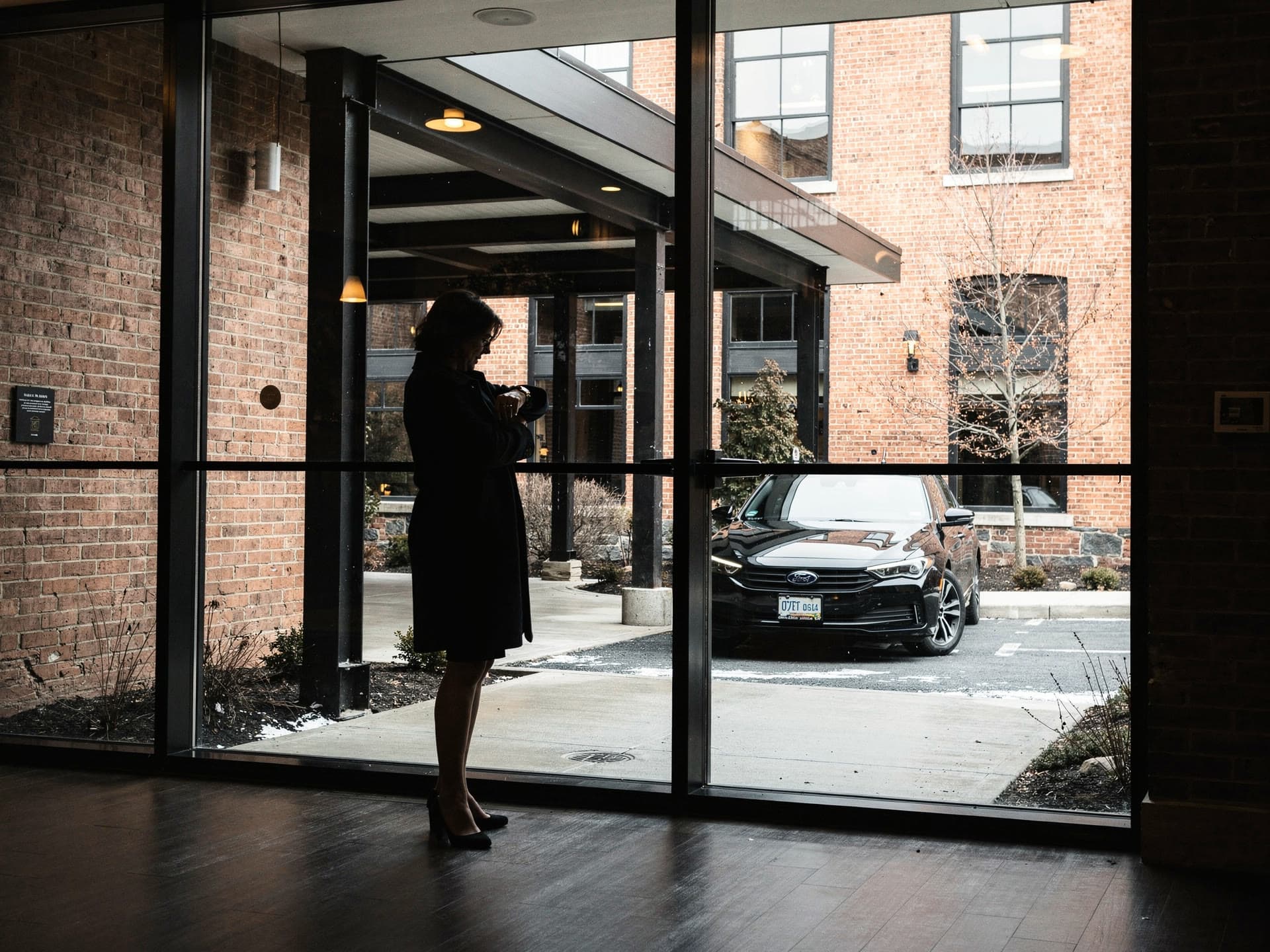 Seen from inside a hotel lobby, a black sedan already waiting under the portico while a woman in a coat checks her watch against the glass.