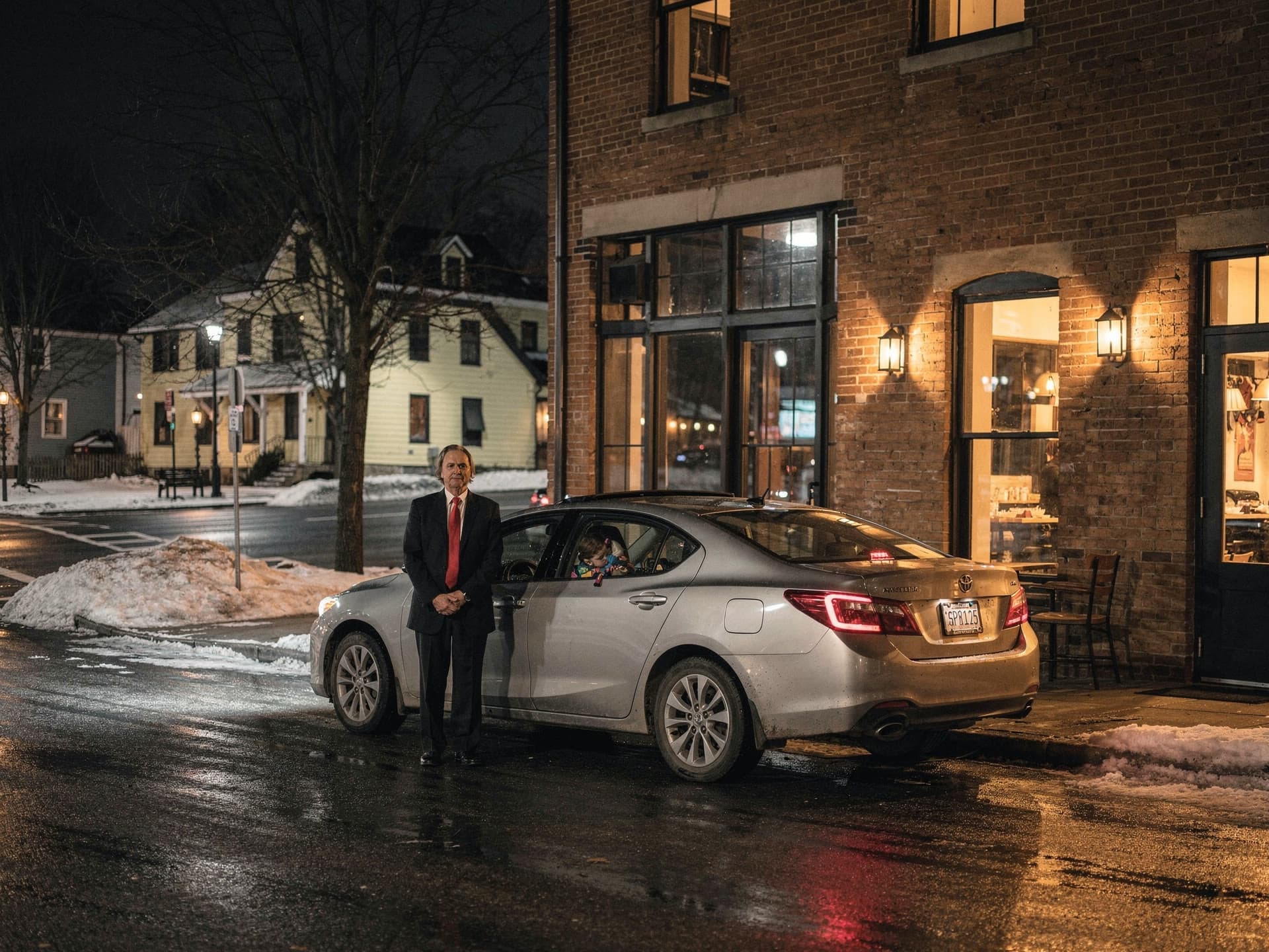 Seen from across the street at night, a chauffeur standing beside an ordinary sedan outside a lit restaurant, waiting.
