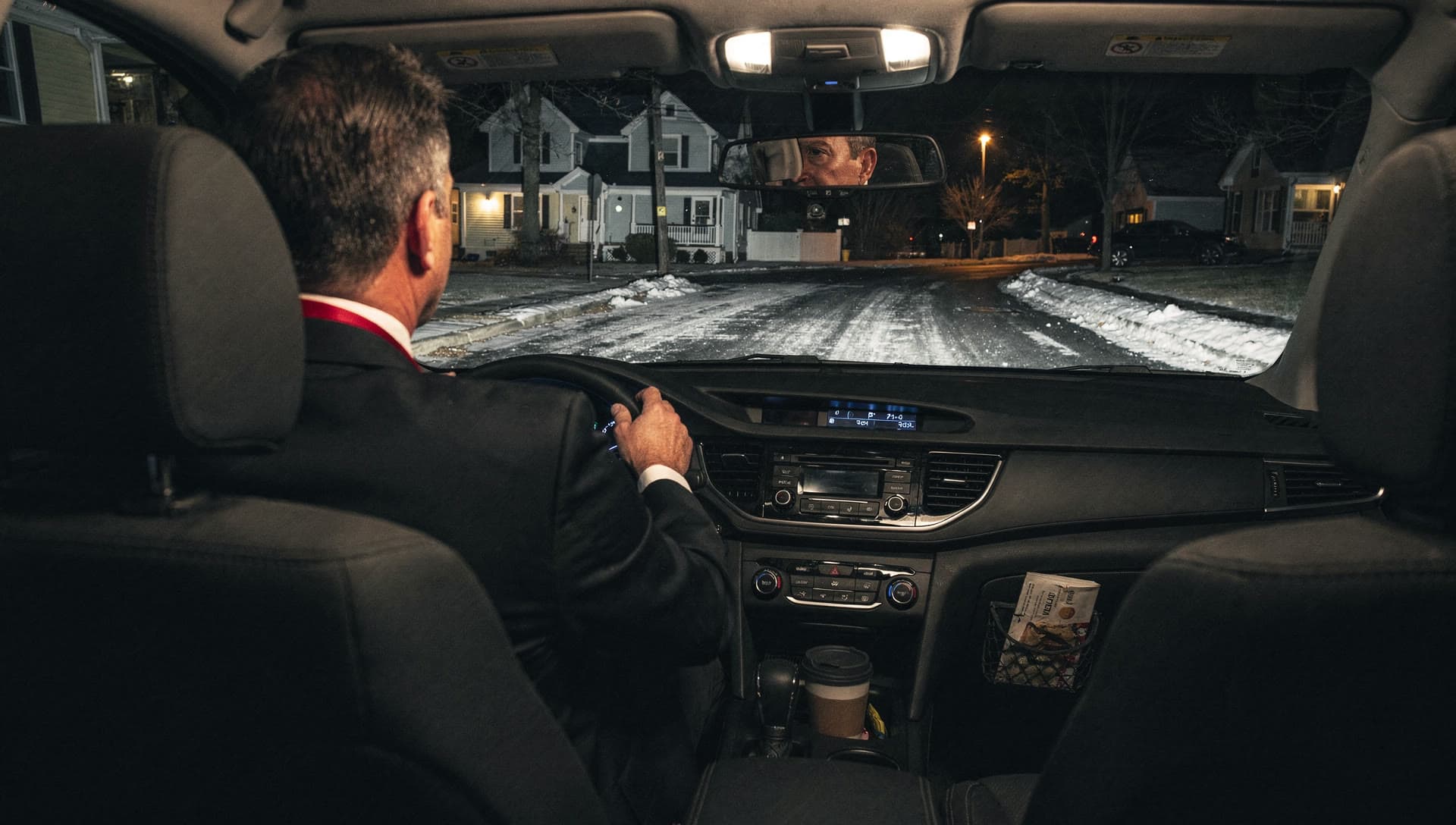 Seen from the rear seat of an ordinary family car at night, a chauffeur's hands on the steering wheel with a quiet suburban street ahead.