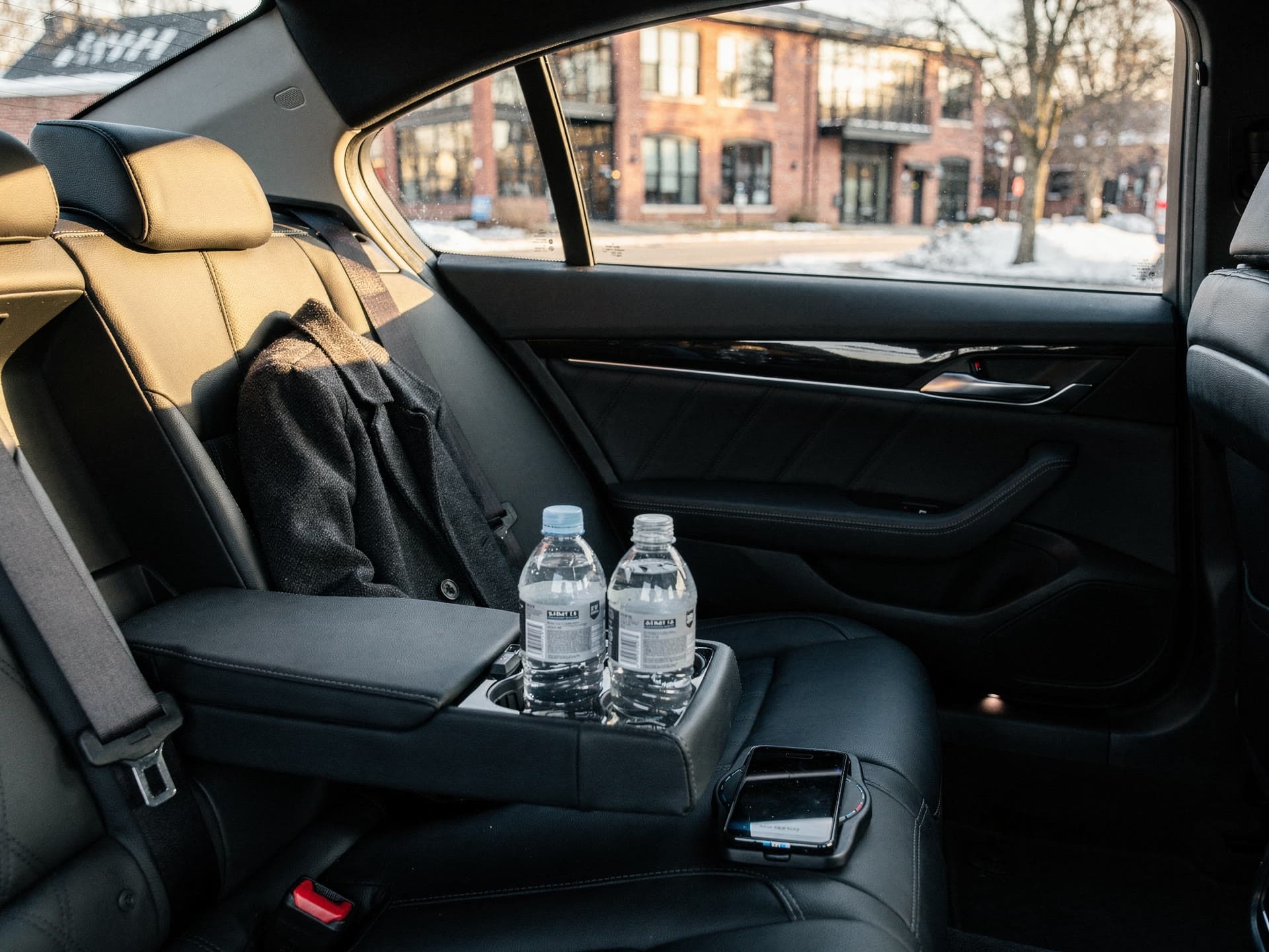 The interior of a waiting sedan in the afternoon, water bottles in the console, a phone on a charger and a folded coat on the seat.