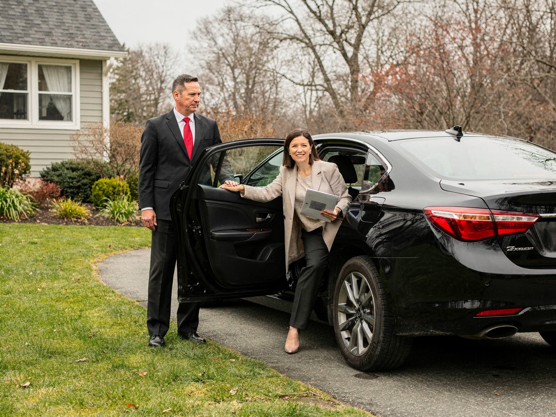 A real estate agent stepping out at a property with a clipboard under her arm while the chauffeur holds the rear door half open.
