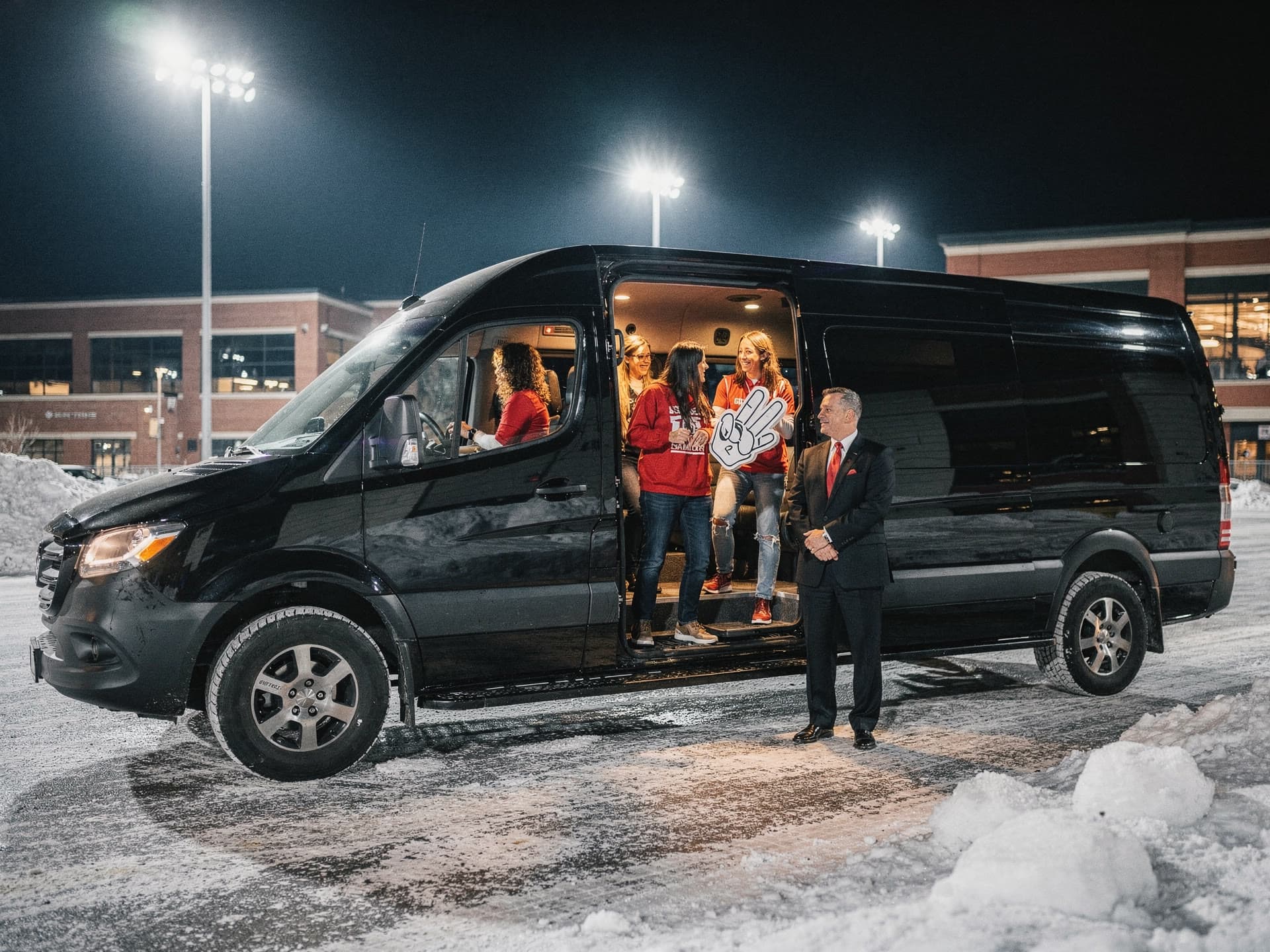 Four friends boarding a Sprinter limo on an arena forecourt after an event, floodlights behind them and one still holding a foam finger.