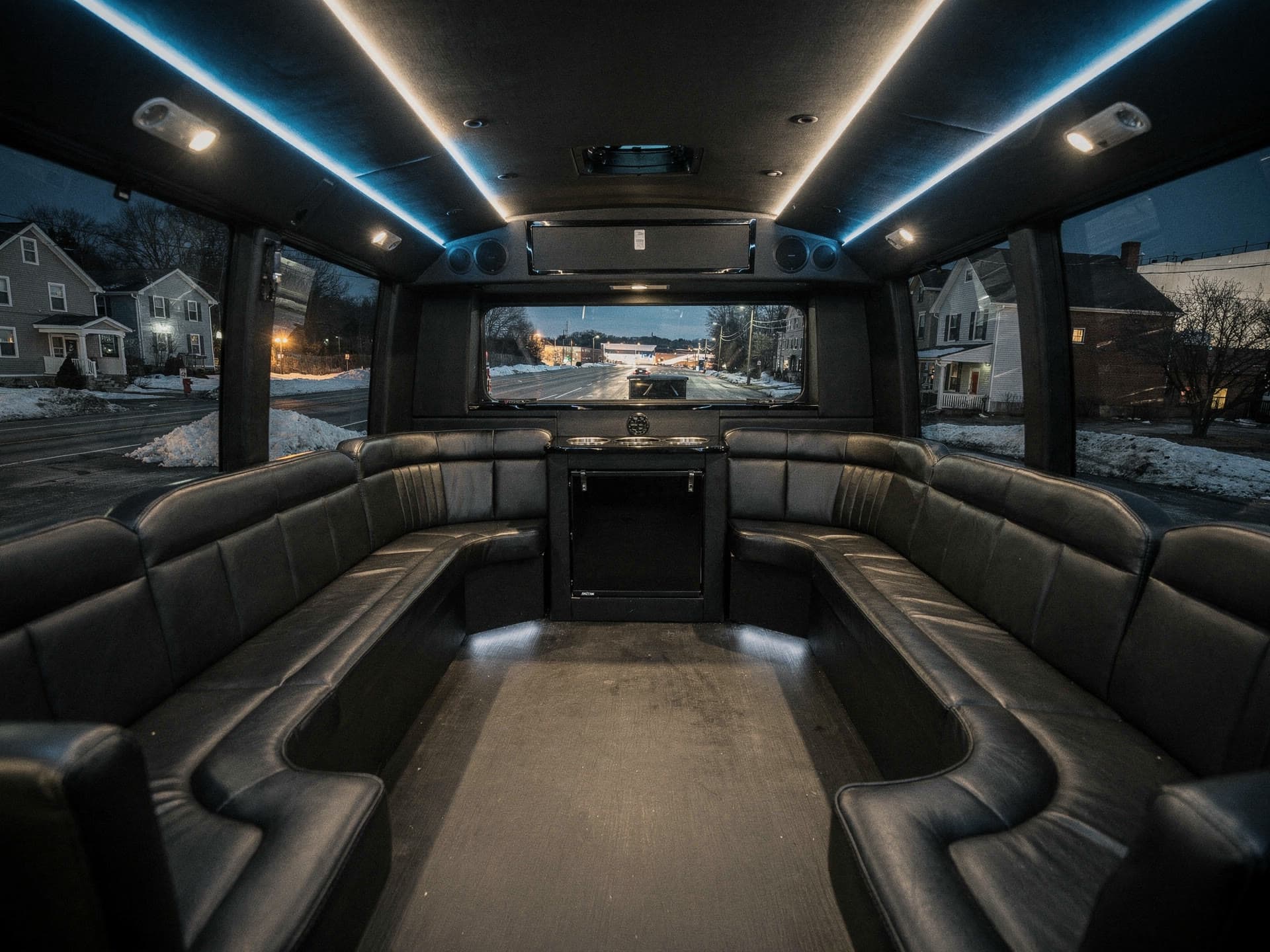The interior of a party bus at night, wraparound leather bench seating lit by coloured ceiling lighting and city glow through tinted windows.