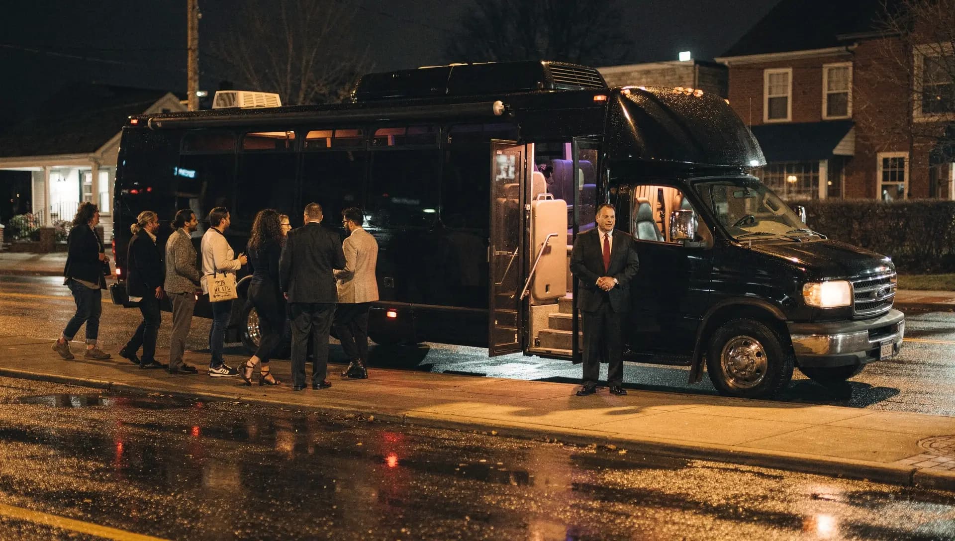 A group walking toward a black party bus at a wet Methuen curb at night, door open and interior lit