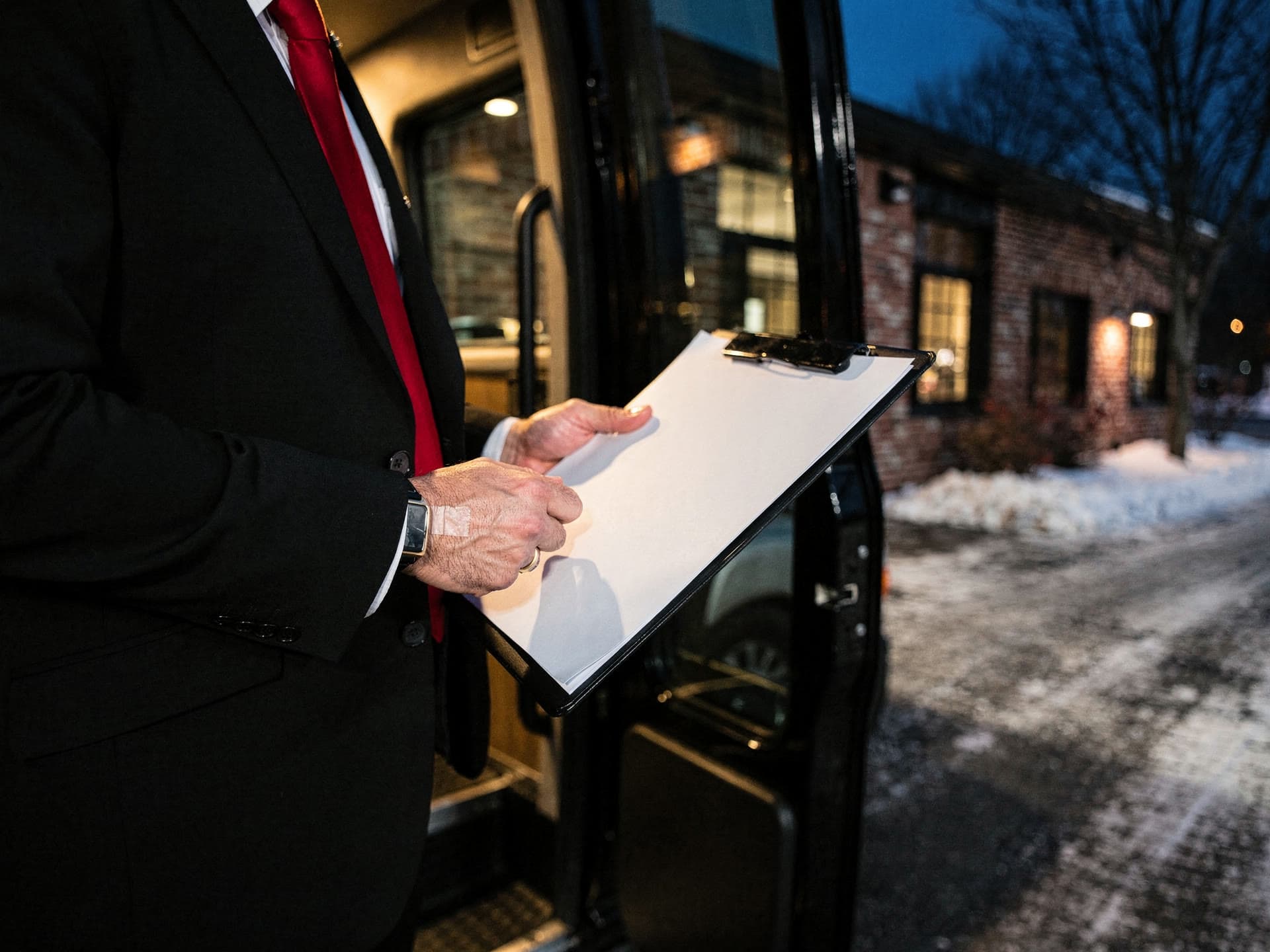 A chauffeur's hands holding a clipboard at the open door of a party bus, red tie entering the frame edge, interior lighting spilling out.