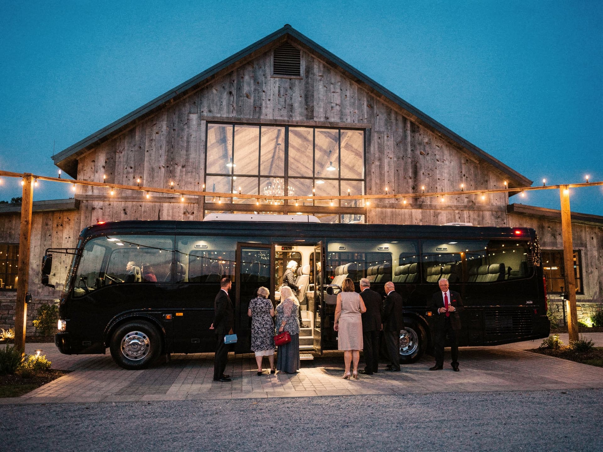 Guests in formal wear boarding a full size coach outside a barn venue at dusk, string lights overhead and a chauffeur counting them aboard.