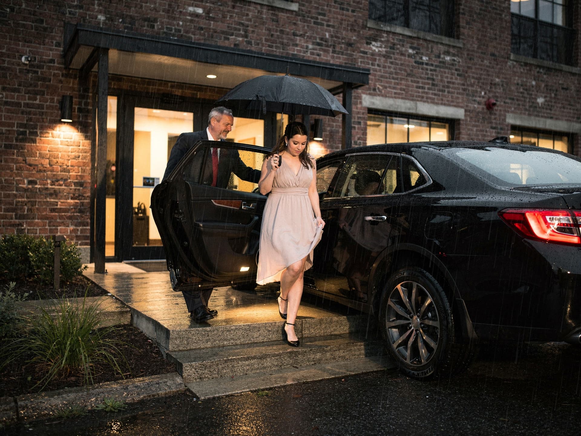 A chauffeur holding a black umbrella over a bridesmaid across three steps of wet stone between a car door and a lit entrance.