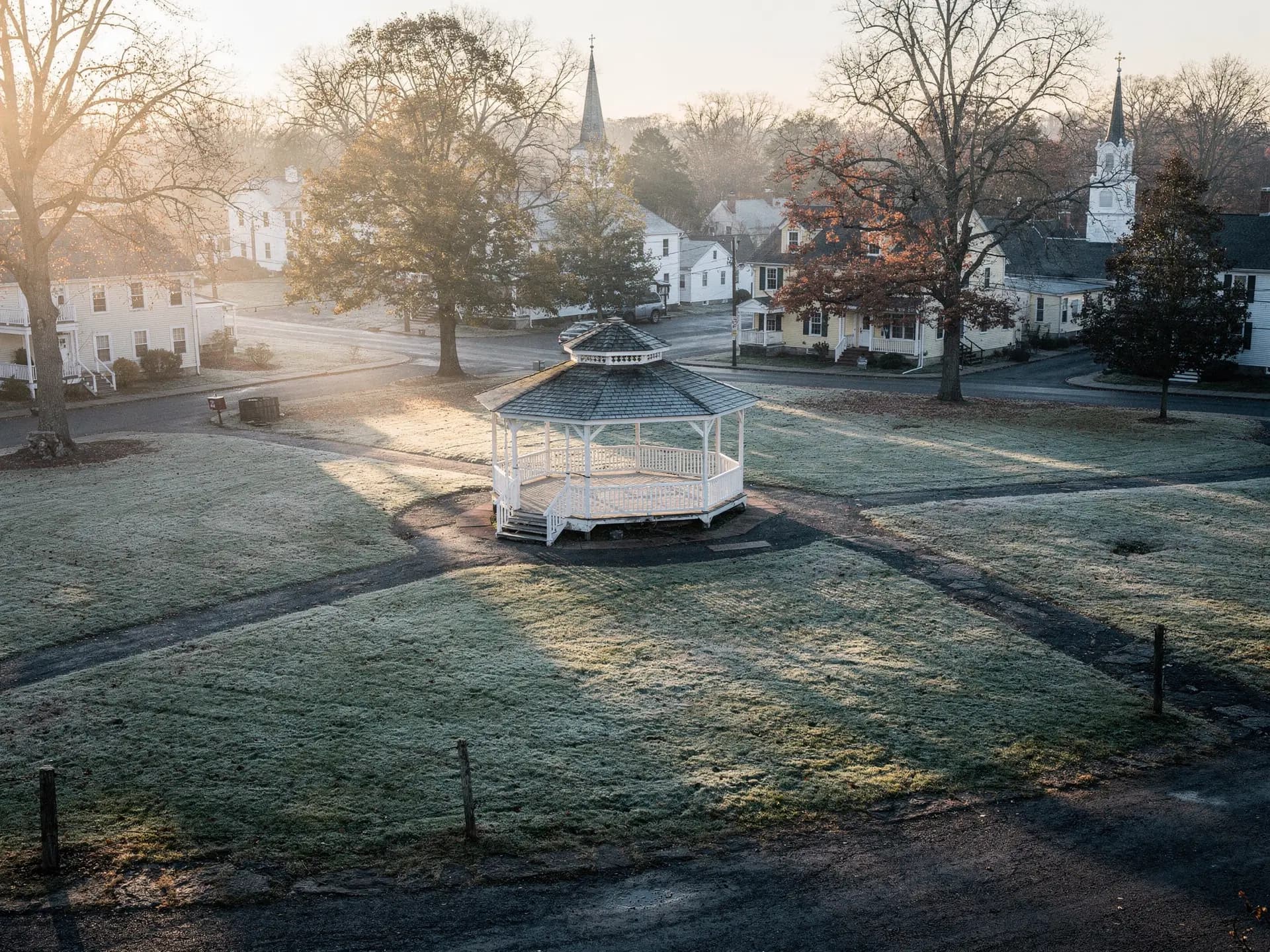 A white bandstand on a village common in early morning light, mist still sitting on the grass around it.
