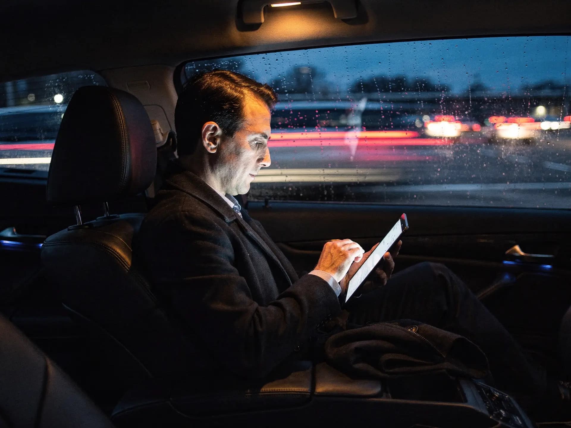 An executive working from a tablet in the rear seat of a sedan at dawn, reading light on, motorway lights passing outside.