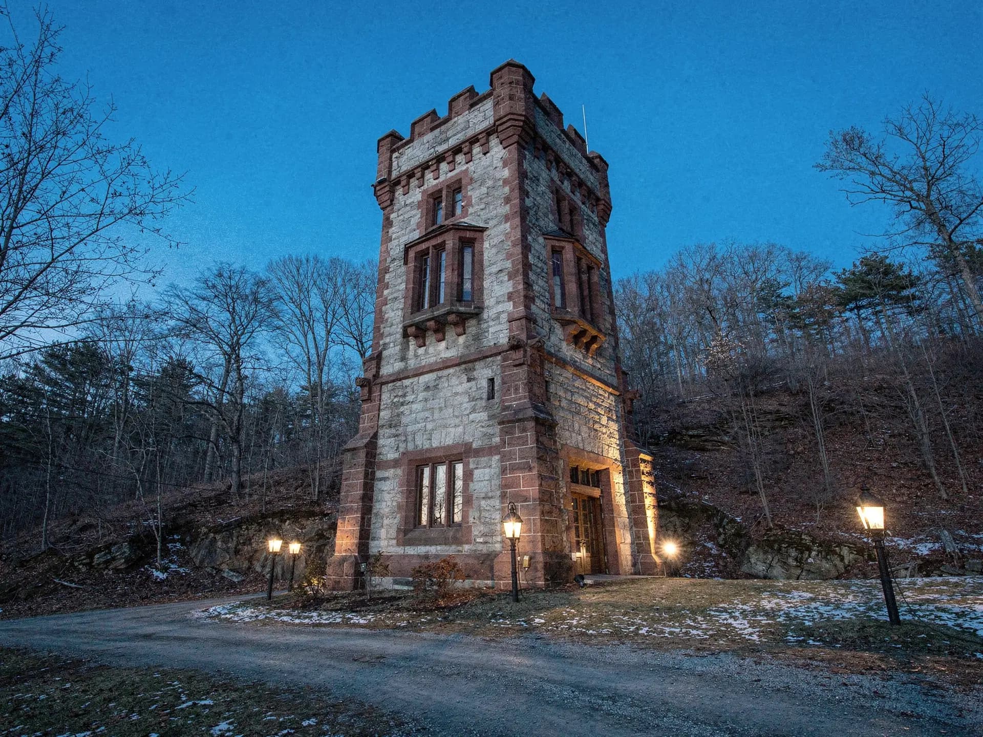 A granite and sandstone Tudor tower rising above bare trees at dusk, lamps lit along the drive below it.