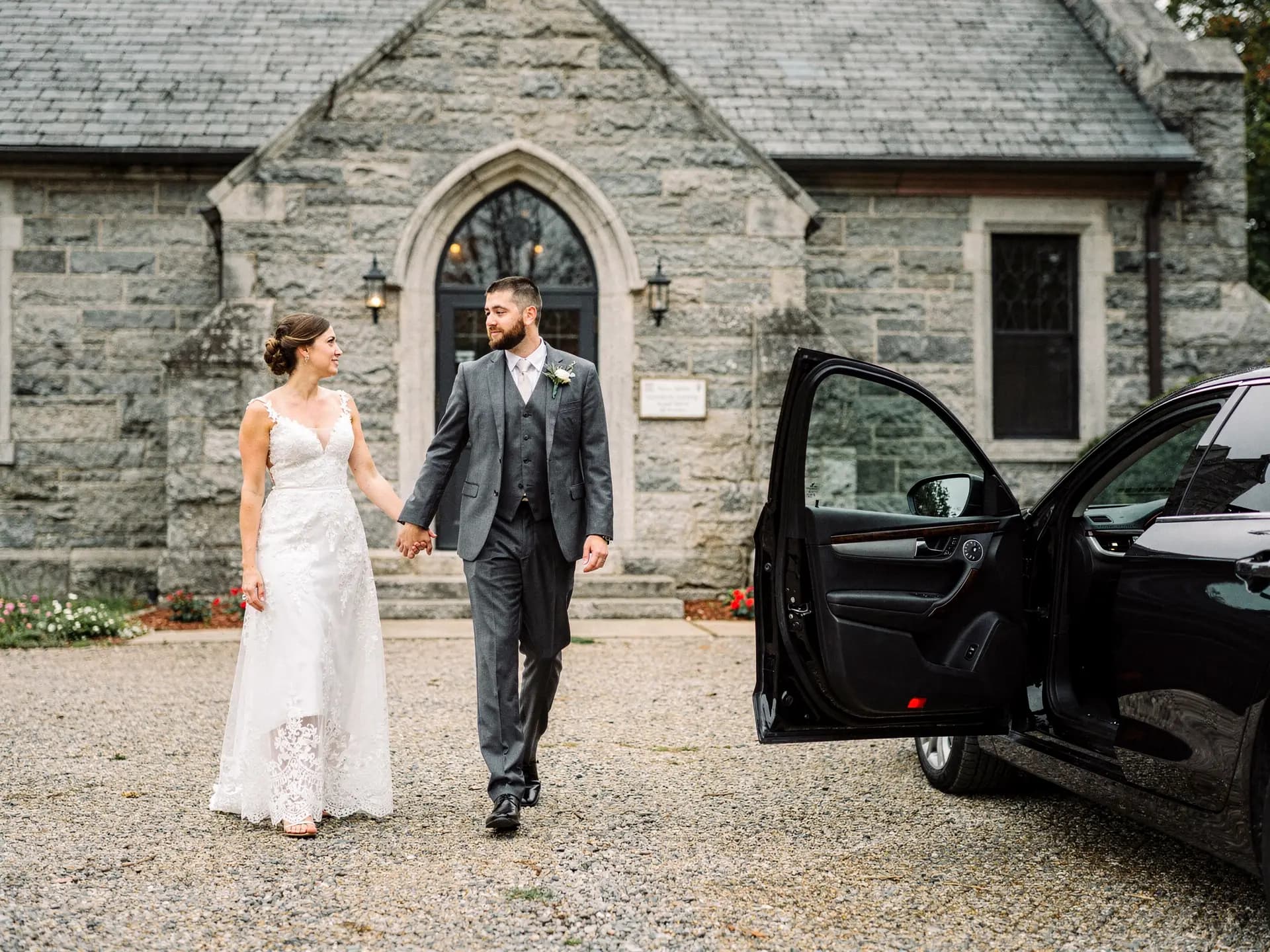 A bride and groom walk hand in hand across the gravel toward a black car with its rear door already open, the stone Gothic chapel behind them.