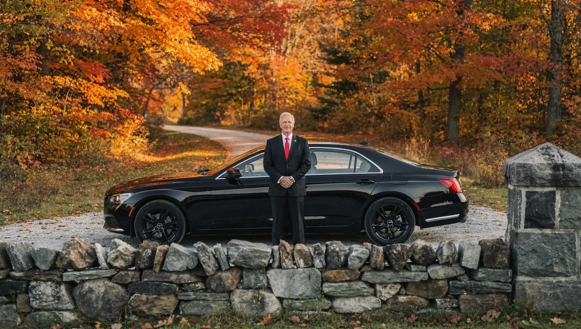 A chauffeur in a black suit stands waiting beside a black car on a Windham lane, a fieldstone wall in front of them and maples in full autumn colour behind.